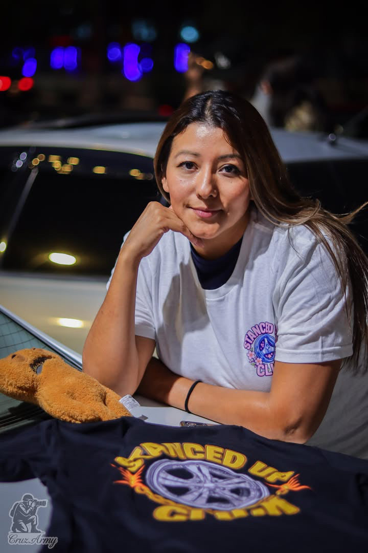 SUC Worldwide On Dubs Tee laid flat on a car hood, black unisex tee with a chrome wheel and flame graphic under gold Stanced Up Click lettering